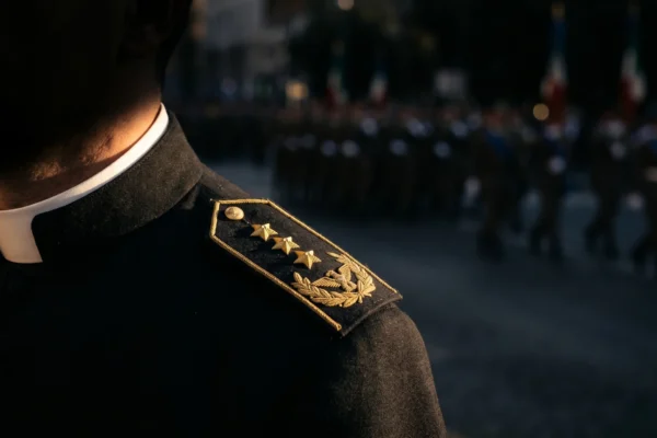 Sacerdote in talare con collarino bianco e spallini militari con stellette dorate, sullo sfondo una parata sfocata con tricolori