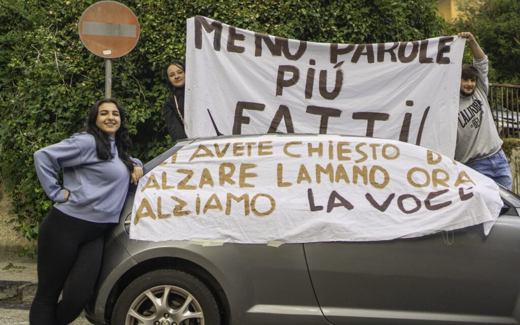 Tre studenti con due striscioni: «Meno parole più fatti» e «Ci avete chiesto di alzare la mano, ora alziamo la voce», uno fissato sulla fiancata di un'auto grigia